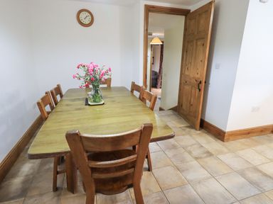 A dining room with a wooden table and six chairs a vase with pink flowers a clock on the wall and an open wooden door at Tithe Cottage in Cockermouth