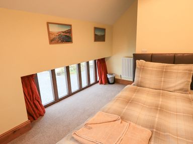 A bedroom with a single bed with beige plaid bedding a row of low windows red curtains and framed pictures on a yellow wall at Tithe Cottage in Cockermouth