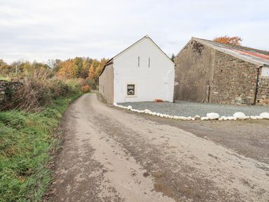 A narrow country road next to a white building and a stone barn with trees in the background at Tithe Cottage in Cockermouth