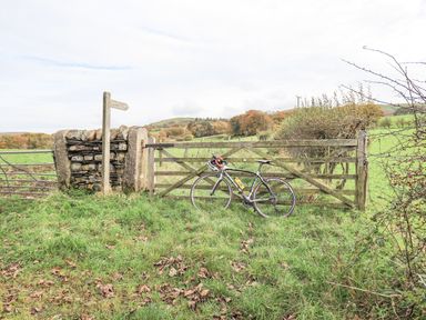 A bicycle leaning against a wooden gate on a grassy path with a stone signpost and fields at Tithe Cottage in Cockermouth