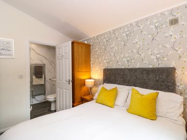 A bedroom with a white bedspread and yellow pillows beside a wooden cabinet and a lamp with floral wallpaper at Footprints Lodge in Windermere