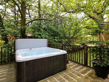 An outdoor wooden deck with a hot tub surrounded by trees and greenery at Footprints Lodge in Windermere