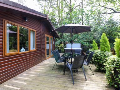 An outdoor wooden deck with a table and six chairs under an umbrella next to a wooden house and surrounded by trees and plants at Footprints Lodge in Windermere