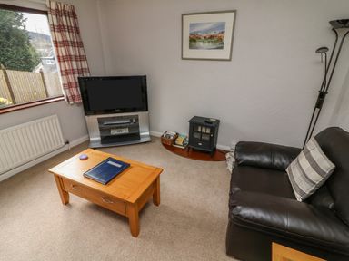 A living room with a brown leather sofa a wooden coffee table a flat screen TV on a stand a basket with books a small stove and a framed picture on the wall at Long Crag Annexe Ambleside