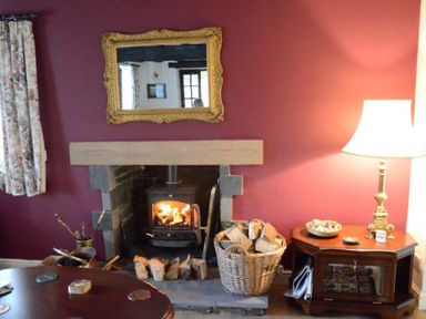 A room with a lit fireplace a basket of firewood a table lamp and a gold framed mirror at Greety Gate House in Foxfield