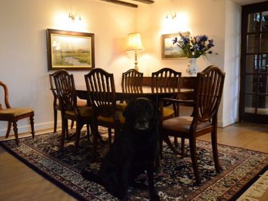 A dining room with a wooden table and chairs a floral rug two framed pictures a lamp a vase with flowers and a black dog at Greety Gate House in Foxfield