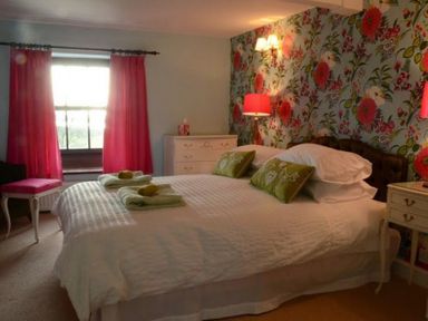 A bedroom with a large bed white bedding floral wallpaper bedside tables and red curtains at Greety Gate House in Foxfield