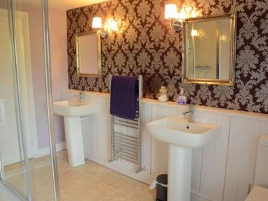 A bathroom with two pedestal sinks two mirrors lights towel rack with purple towel and floral wallpaper at Greety Gate House in Foxfield