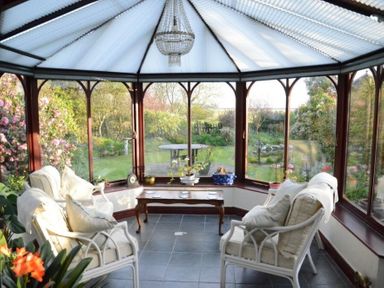 A conservatory with cushioned armchairs a wooden coffee table and a chandelier overlooking a garden at Greety Gate House in Foxfield