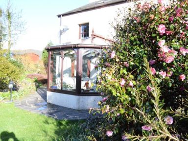 A garden with a stone path leading to a conservatory attached to a house with pink flowers on a bush at Greety Gate House in Foxfield