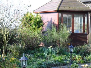 A garden with various plants and lamp posts in front of a wooden building with windows at Greety Gate House in Foxfield