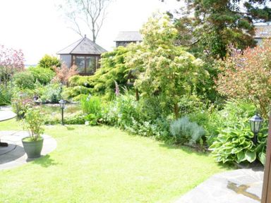 A garden with green plants bushes and a pond with a small glass gazebo in the background at Greety Gate House in Foxfield
