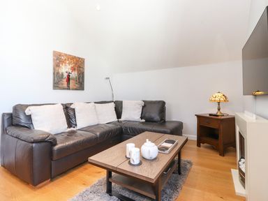 A living room with a black leather sectional sofa with white cushions a wooden coffee table with a teapot and candles a side table with a lamp and a wall-mounted tv at Sky View in Keswick