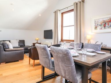 A dining area with a wooden table set for six and grey chairs next to a living room with dark sofas and a wall-mounted TV at Sky View in Keswick