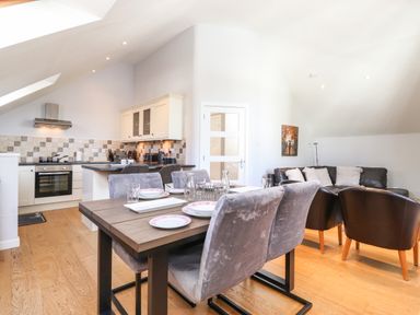 An open dining and living area with a wooden dining table set with plates and glasses a kitchen with an oven and cabinets and a seating area with sofas and armchairs at Sky View in Keswick