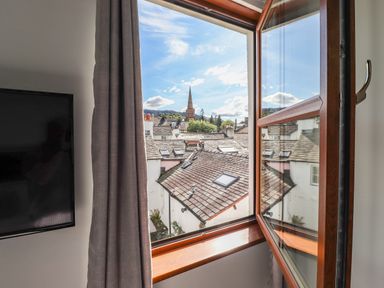 View through an open window showing rooftops and a church steeple with a TV and curtain inside the room at Sky View in Keswick