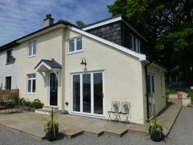 A yellow two-story house with black door and white framed windows a stone patio with two black chairs and potted plants at Eribel Cottage in Threlkeld