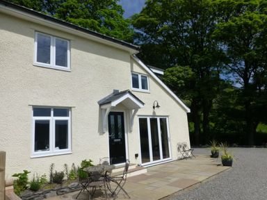 An exterior view of a cream colored house with black door and white windows patio furniture and plants on a stone patio at Eribel Cottage in Threlkeld