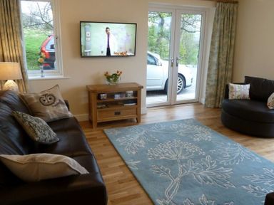 A living room with two sofas a wooden TV stand a wall-mounted TV and large windows at Eribel Cottage in Threlkeld