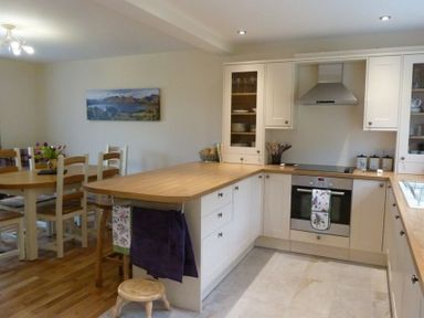 A kitchen with white cabinets wooden countertops a built in oven and stovetop and a dining area with a wooden table chairs and a vase with flowers at Eribel Cottage in Threlkeld