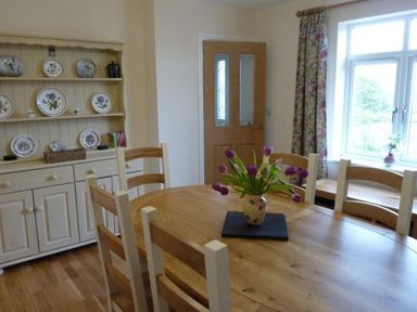 A dining room with a wooden table and chairs a vase with flowers on the table a cupboard with decorative plates and a window with floral curtains at Eribel Cottage in Threlkeld