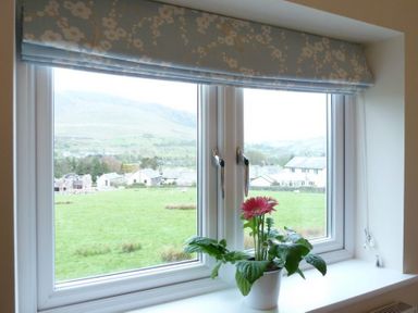 A window with a floral roller blind and a potted plant on the sill overlooking a green field and houses at Eribel Cottage in Threlkeld