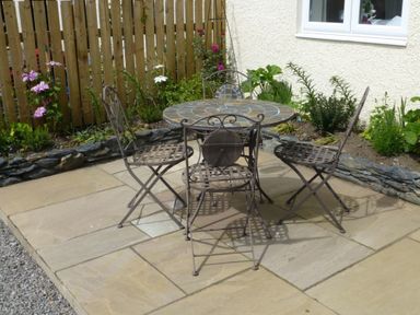 An outdoor patio with a round table and four metal chairs on stone tiles next to a garden bed and wooden fence at Eribel Cottage in Threlkeld