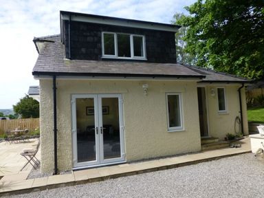 Exterior view of a two-story house with white framed windows and doors a patio with small table and chairs and gravel driveway at Eribel Cottage in Threlkeld