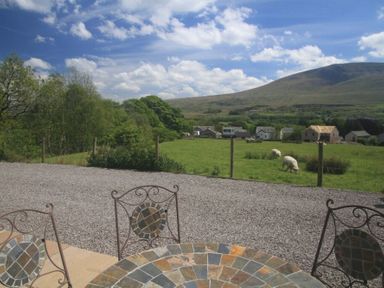 Outdoor patio with stone table and metal chairs overlooking a fenced green field with sheep trees and houses and a hill in the background at Eribel Cottage in Threlkeld