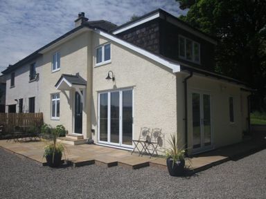 The exterior of a two-story house with a gravel driveway and a paved patio area with outdoor furniture and potted plants at Eribel Cottage in Threlkeld