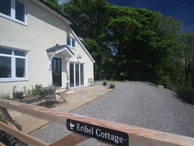 The exterior of a white cottage with outdoor metal chairs and table on a stone patio and a gravel driveway surrounded by trees at Eribel Cottage in Threlkeld