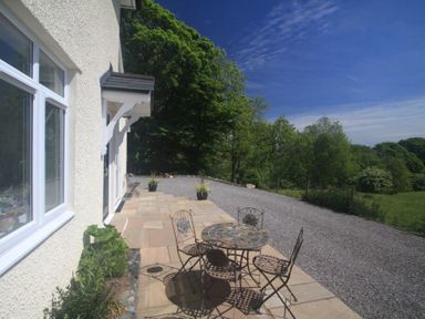 A patio with a round table and four chairs outside a white house with trees and greenery in the background at Eribel Cottage in Threlkeld