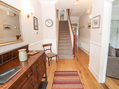 A hallway with wooden flooring a carpeted staircase a wooden sideboard with a ceramic vase and a pen on it a wooden chair a wall clock and framed pictures at White House in Keswick