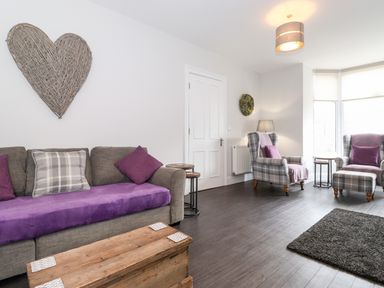 A living room with a grey sofa covered with a purple throw and cushions a wooden chest as a coffee table two plaid armchairs with purple cushions near a window at Latrigg House in Keswick
