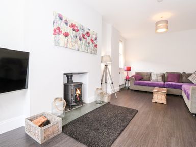 A living room with a wall-mounted television a wood-burning stove a grey sectional sofa with purple cushions and blankets wooden floor and a floral painting above the stove at Latrigg House in Keswick