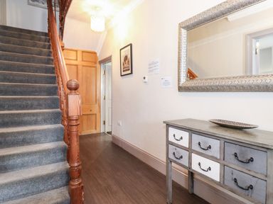 An entryway with a wooden staircase carpeted in grey a wooden door a console table with drawers and a large wall mirror at Latrigg House in Keswick