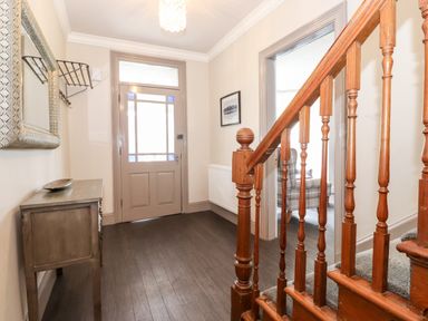 A hallway with a wooden staircase a console table with a dish a wall mirror a door with glass panels and an adjoining room with a chair at Latrigg House in Keswick