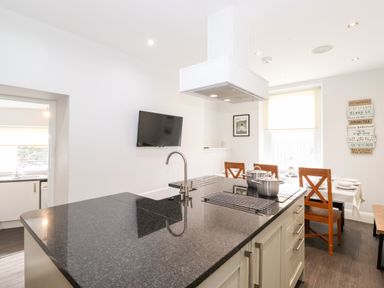 A kitchen with a black countertop island stove and sink with pots a dining table with chairs and a wall mounted tv at Latrigg House in Keswick