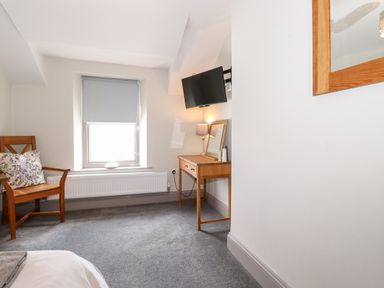 A room with a wooden chair with floral cushion a window with a blind a wooden desk with a table lamp and a wall-mounted TV at Latrigg House Keswick