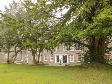 A stone building with multiple windows behind large trees and a grassy lawn at Derwentwater Apartment in Keswick
