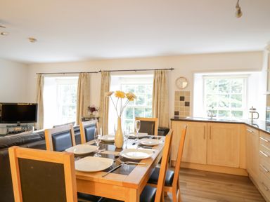 A dining area with a wooden table set with plates and glasses next to a kitchen with light wood cabinets and windows at Derwentwater Apartment Keswick