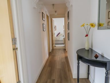 A hallway with wooden flooring a black half-moon table with a vase of flowers and framed pictures on the walls at Derwentwater Apartment Keswick