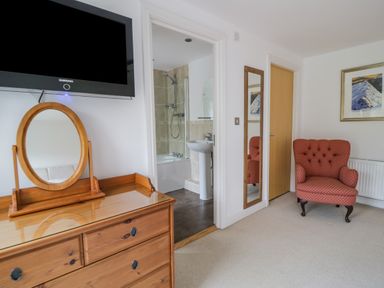 A bedroom with a wooden dresser and mirror a wall-mounted television a red patterned armchair a framed mountain picture a full-length mirror and a bathroom entrance at Derwentwater Apartment in Keswick