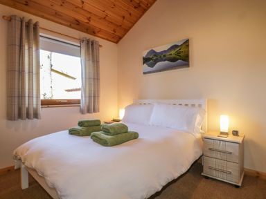 A bedroom with a double bed with white linens and green towels on the bed a nightstand with a lamp and clock and a window with curtains at Holmedell in Greystoke