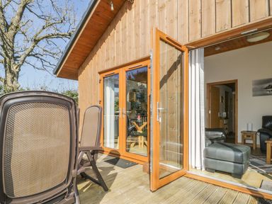An outdoor wooden deck with plastic chairs and an open wooden framed glass door leading to a living room with black leather furniture at Holmedell in Greystoke