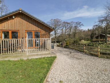 A wooden cabin with a deck and outdoor furniture next to a gravel path and trees at Holmedell in Greystoke