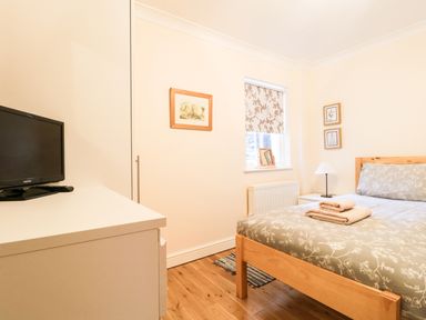 A bedroom with a wooden bed with folded towels a bedside table with a lamp a window with patterned blind a white dresser with a TV on top and framed pictures on the walls at Siskin in Keswick