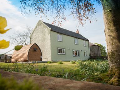 A two-story house with green walls and a small wooden curved shed in a garden with trees and flowers at Westmains Farm in Penton