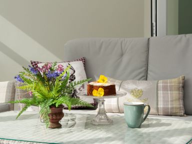 A glass table with a flower arrangement a cake on a stand and a mug in front of a sofa with cushions at Westmains Farm in Penton