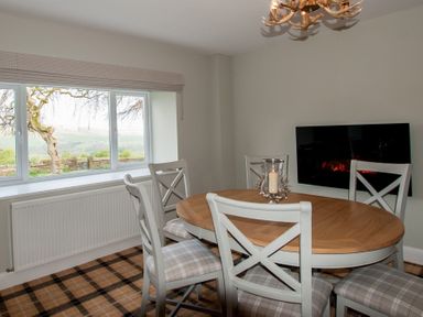 A dining room with a wooden oval table surrounded by white chairs with plaid cushions and a window showing a tree and hills at Westmains Farm in Penton